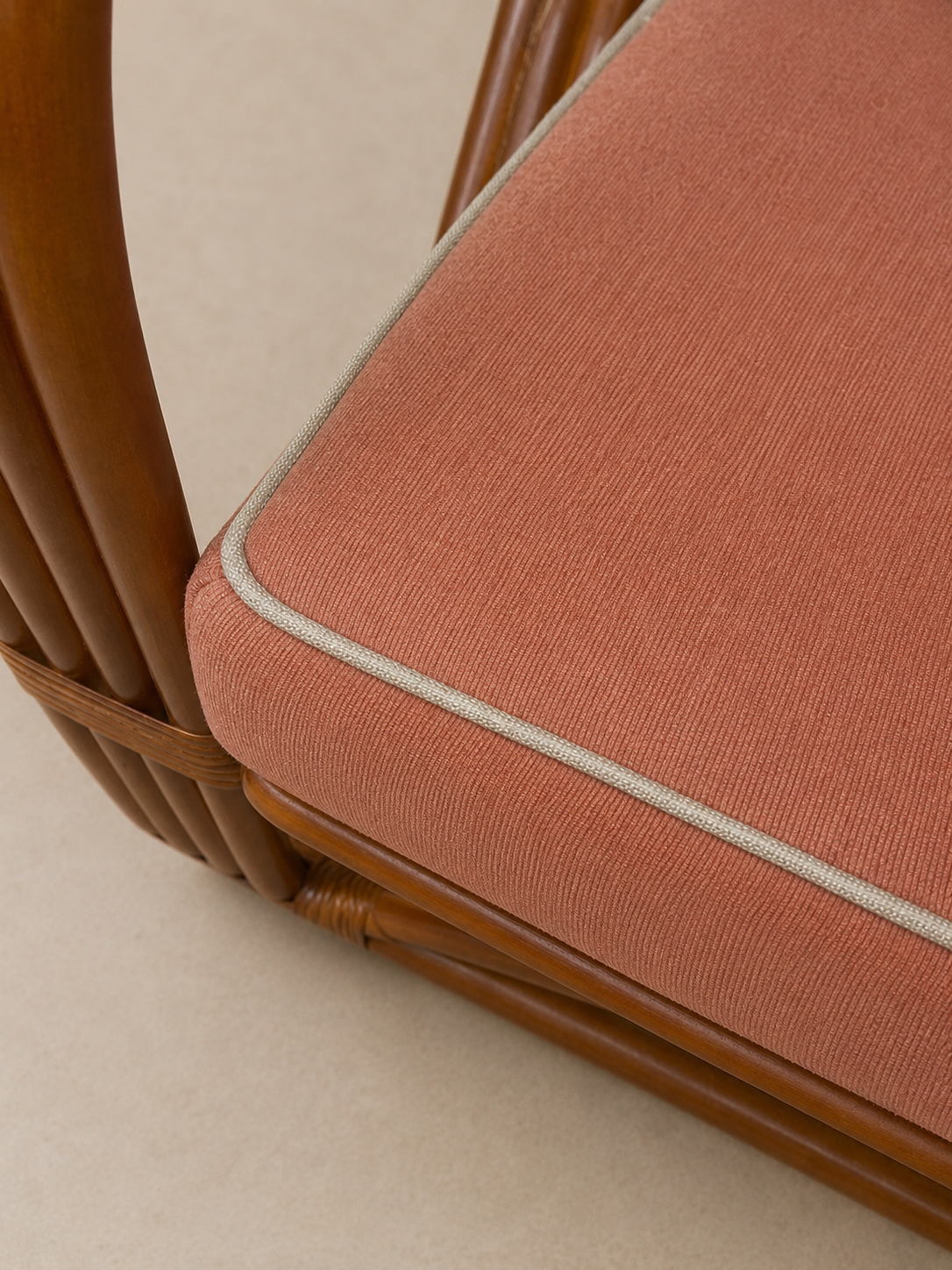 Close-up of a woven rattan chair with a textured dusty rose cushion and light beige piping, set against a neutral beige background.