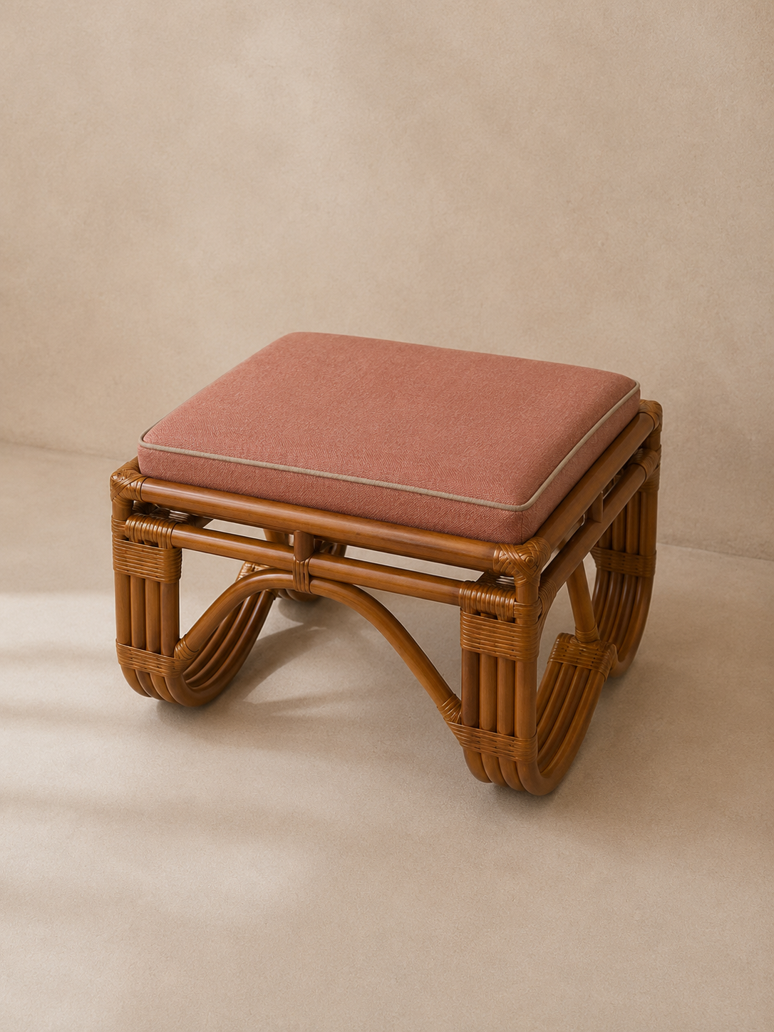A small woven rattan stool with a square dusty rose cushion, photographed against a neutral beige background.